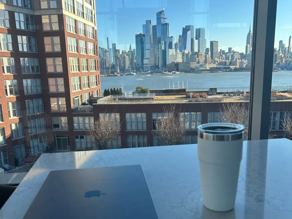 Chris's laptop and coffee on a marble table in the Maxwell Place lounge, with the Hudson River and the Manhattan skyline outside the window
